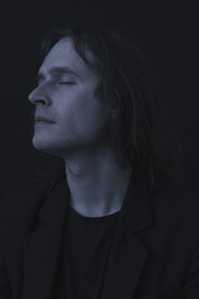 Somber portrait of Błażej, sharp edges, delicate blue light. His eyes are closed.